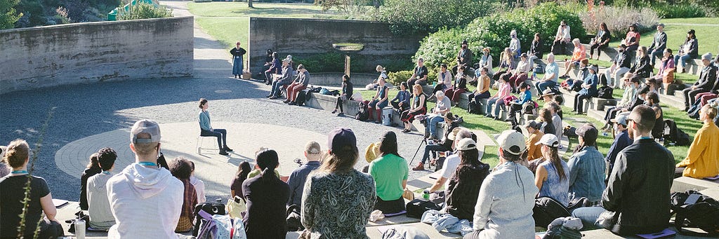 A crowd of people sit in an outdoor amphitheatre surrounded by grass and shrubs. One person sits in a chair in the middle of the stage as the crowd looks on.