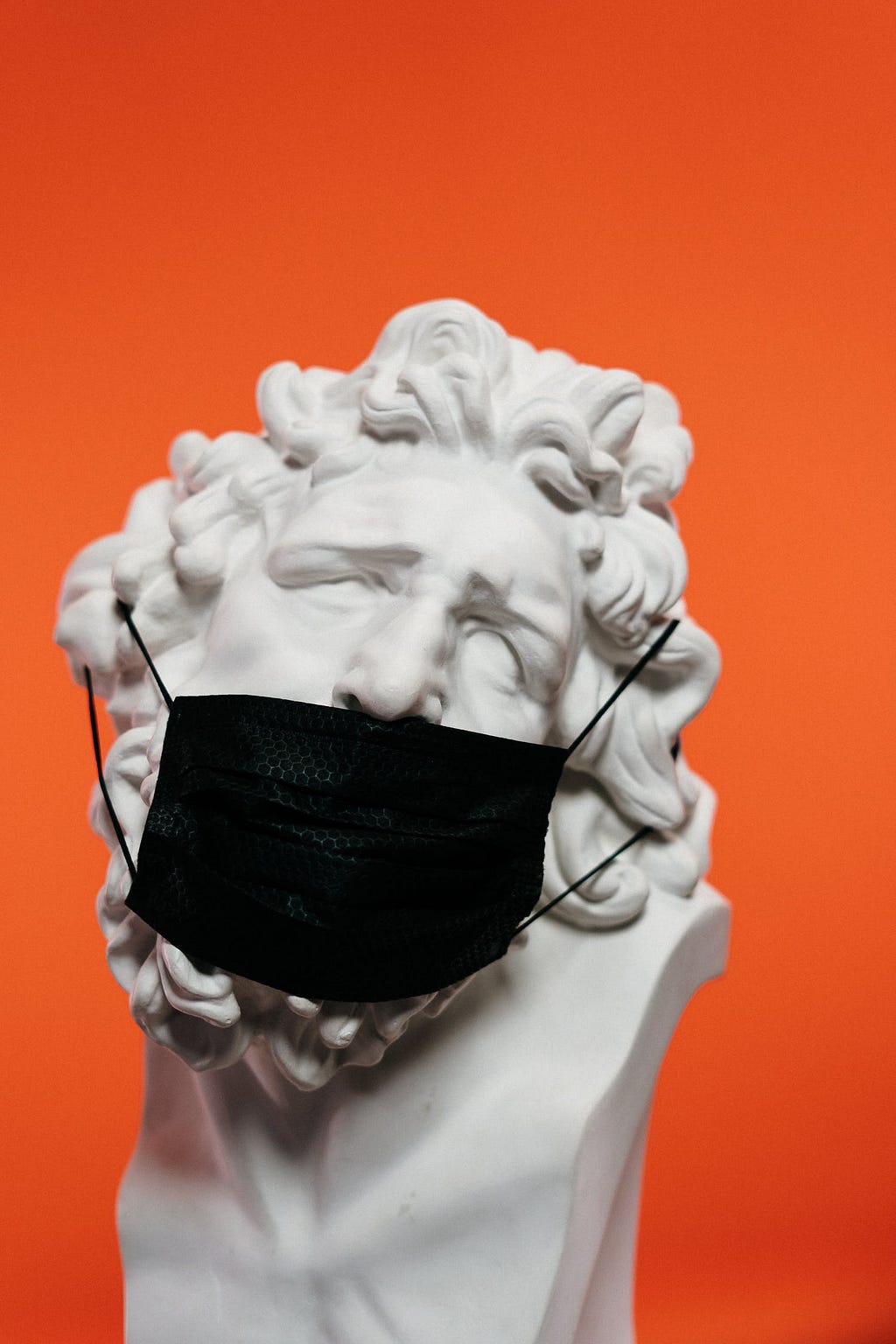 An ancient greek statue with a black medical mask in an orange background.