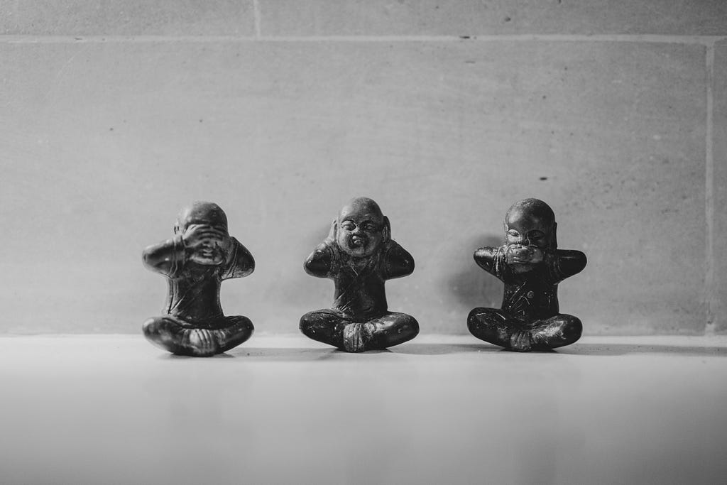 Three bald-headed statuettes sit crosslegged on the floor. From left to right they use their hands to close their eyes, ears, and mouth.