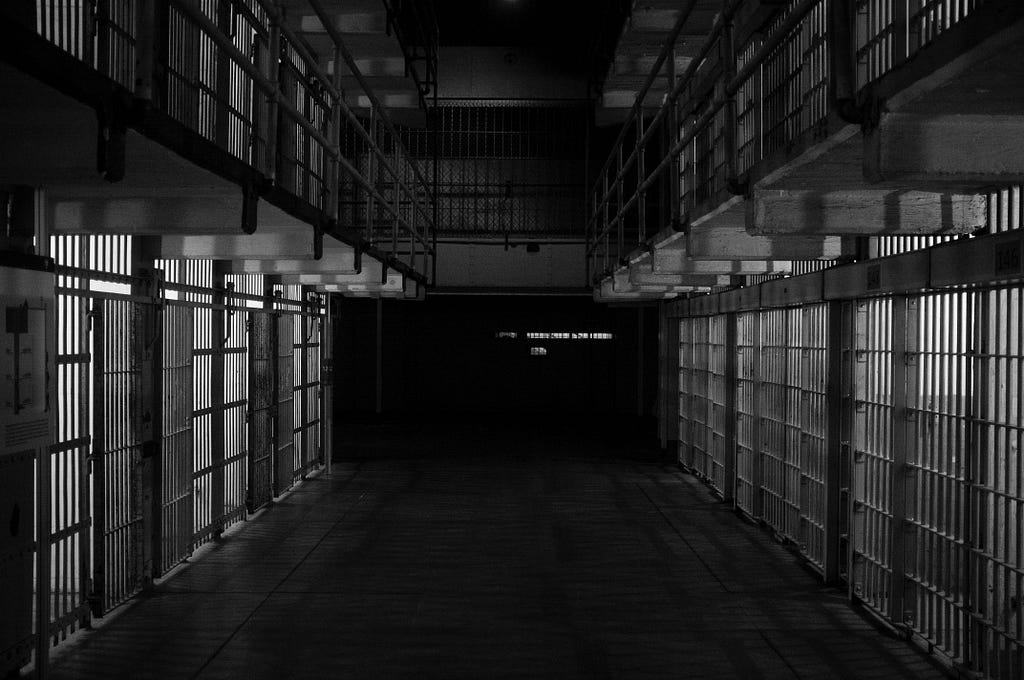 A black-and-white photograph of a corridor within a prison, closed cell gates on either side.