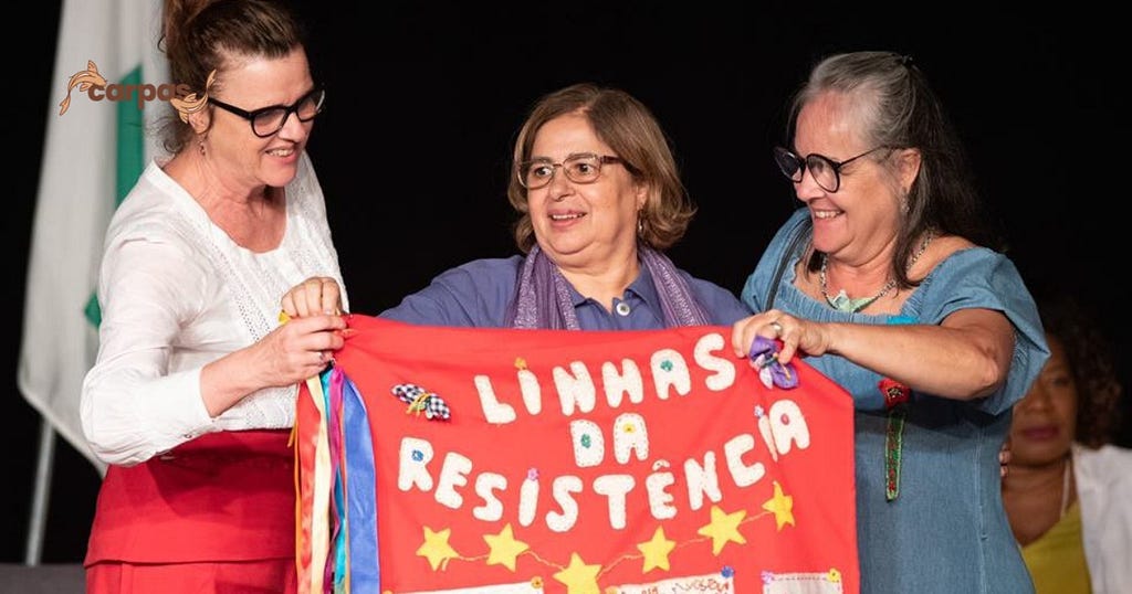 Na foto, a ministra das Mulheres Cida Gonçalves aparece ao centro segurando um tecido vermelho com os dizeres “LINHAS DA RESISTÊNCIA” em letras brancas e seis estrelas amarelas embaixo. Ao seu lado estão duas mulheres, companheiras de luta da ministra.