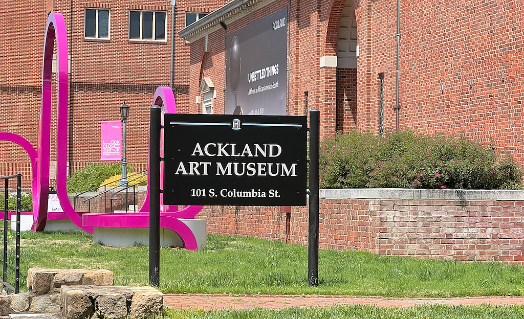 A picture of the Ackland Art Museum. The building is brick with grass ans shrubs around. There is a pink sculpture in front and a sign that reads Ackland Art Museum 101 S. Columbia St.