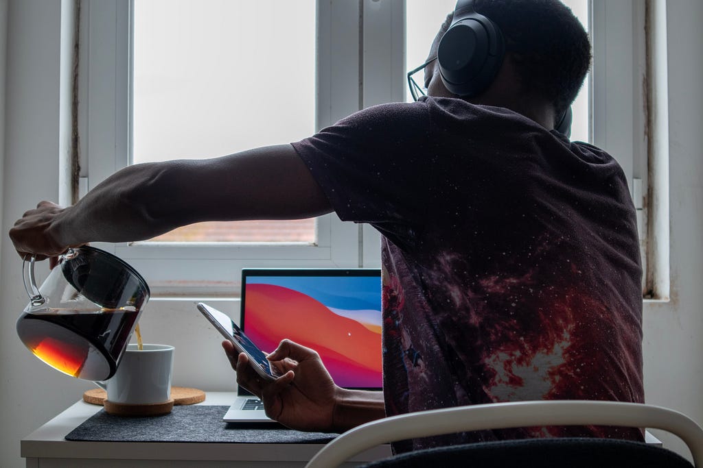 a person sits in front of a window. their laptop is on the table in front of them, they are wearing headphones, a phone is in their right hand and they are pouring tea into a mug with their left hand.