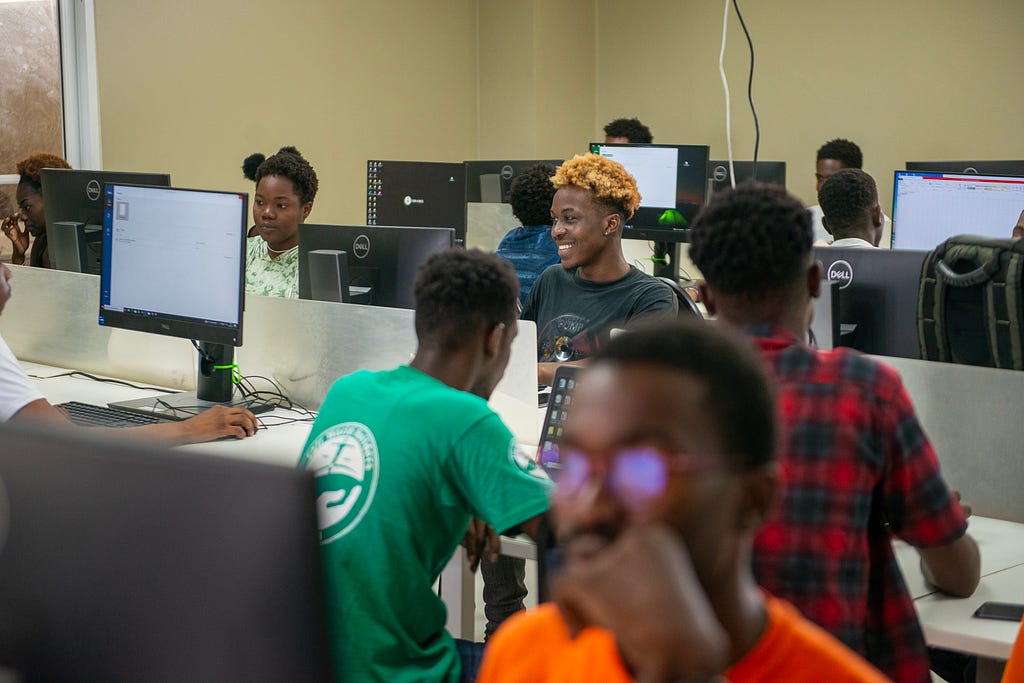A group of students sit at computer monitors in a classroom in Haiti designed for virtual learning.