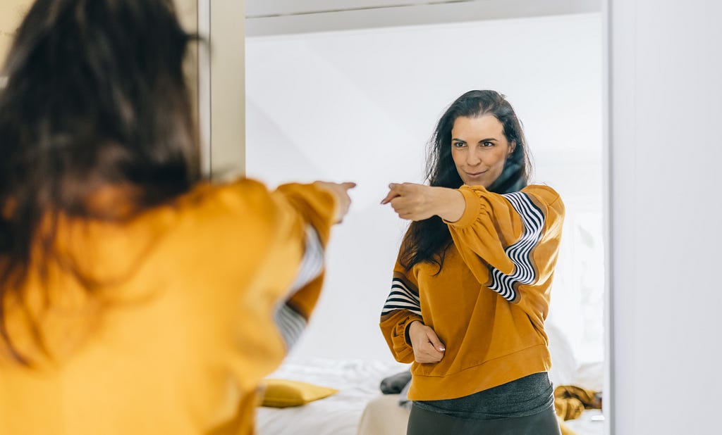 A girl in a yellow sweatshirt points to herself in the mirror while smiling
