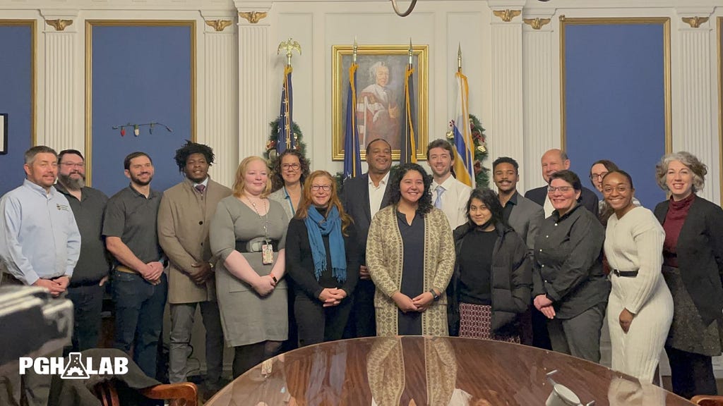 17 members of city government and companies standing, smiling, and posing together in a decorated city conference room