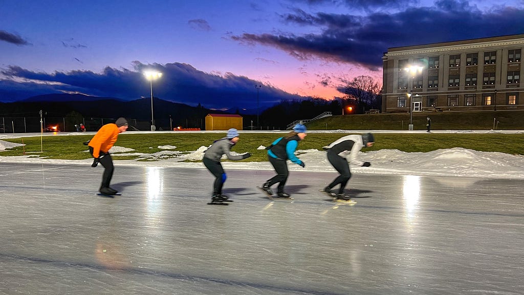 Photo of World Ice Skating Day participants skating on the outdoor Olympic Speed Skating Oval with a blue and pink sunset off in the distance.