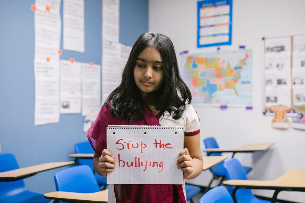 Indian girl in a classroom wearing a pink and white sari, holding up an open notebook with the words “stop the bullying” written across the page in red marker