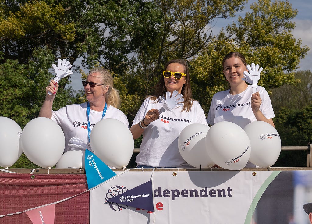 Independent Age staff wear charity t-shirts while smiling and waving at the side of a running route. They have balloons and an Independent Age banner to cheer on runners.