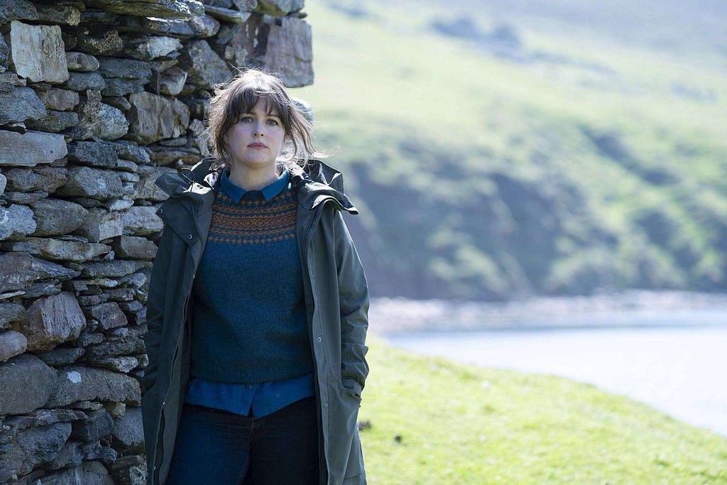 A brunette woman in a blue jumper and long green coat stands next to a dry stone building next to the coast.