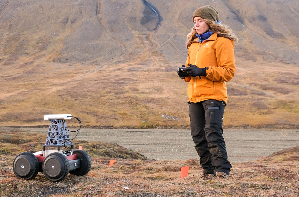 Cody Paige in an orange winter jacket holds a remote controller to direct and test a four-wheeled rover from the MIT Resource Exploration and Science of our Cosmic Environment (RESOURCE) project.