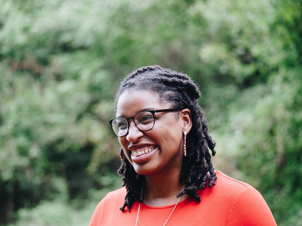 Mulher negra sorrindo, de tranças, com um óculos de grau preto.