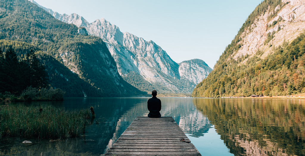 a person sitting on a wooden bridge at a crystal clear lake surrounded by mountains, meditating