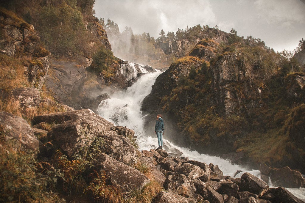 woman standing in front of a waterfall with forest in the background in moody weather