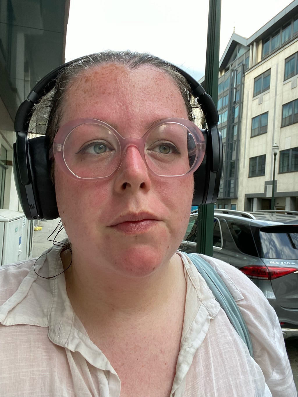 A photo of Bella’s face which is red and blotchy. She is wearing large glasses and black headphones, the background is a urban street.