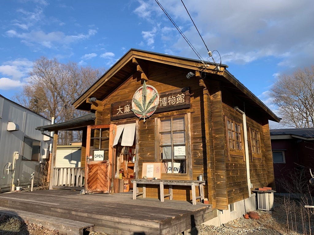 A small wooden one-storey building with a pointed roof and a bench outside. A large sign with a cannabis leaf and the name of the museum in Japanese is on the front of the building.