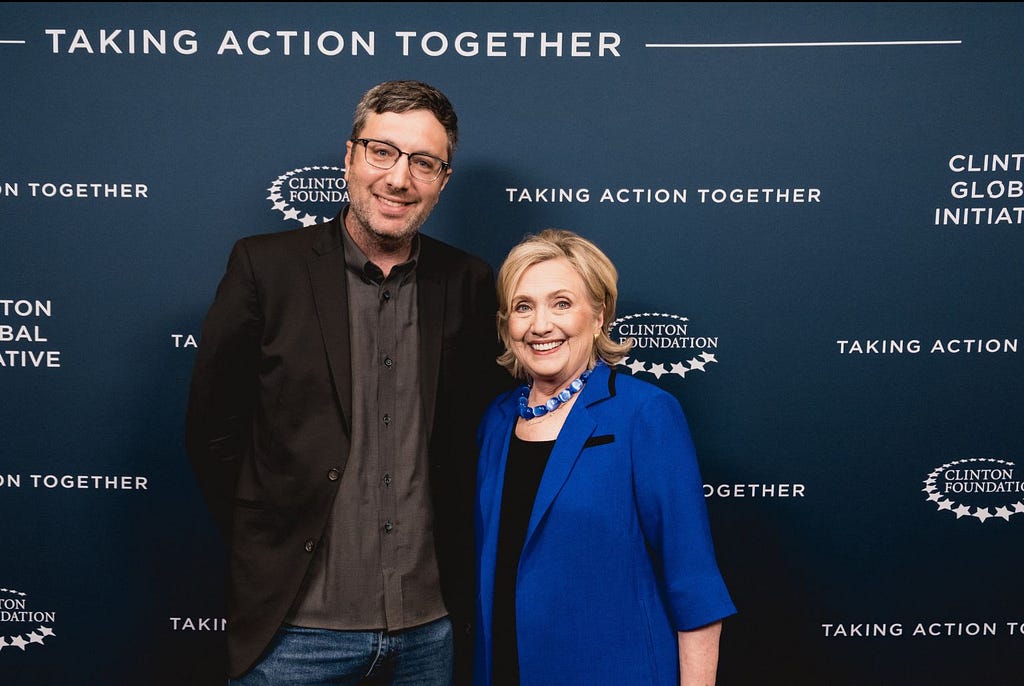 Sostento CEO Joe Agoada wearing a dark jacket standing next to former Secretary of State Hillary Clinton wearing a blue jacket. Both are smiling at the camera behind a navy background with the CGI logo.