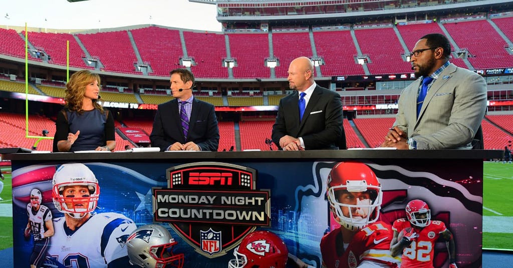 A photo of ESPN’s Monday Night Football panel, featuring four panelists talking with each other, in front of an empty stadium. The banner in front of their desk shows Tom Brady of the New England Patriots on the left and Alex Smith of the Kansas City Chiefs on the right.