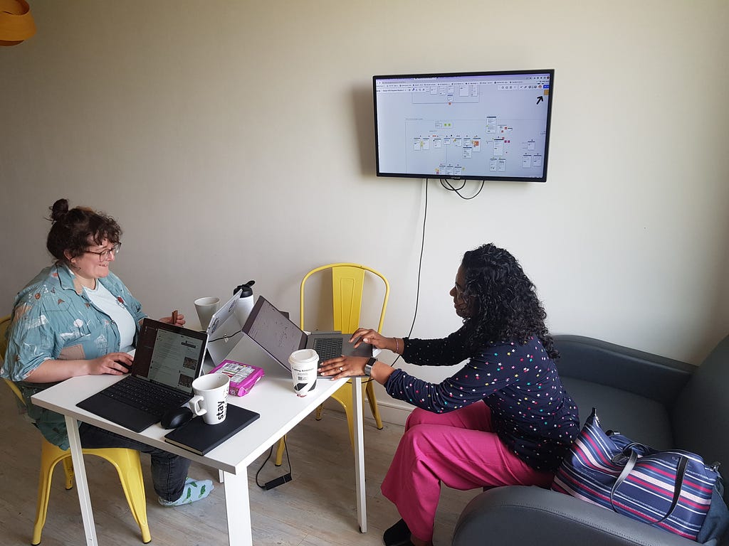 A picture of our work setup. There is a table with 3 laptops on it; 2 people are sat around the table working on their laptops. On the wall there is a tv screen, which is presenting images from one of the laptops.