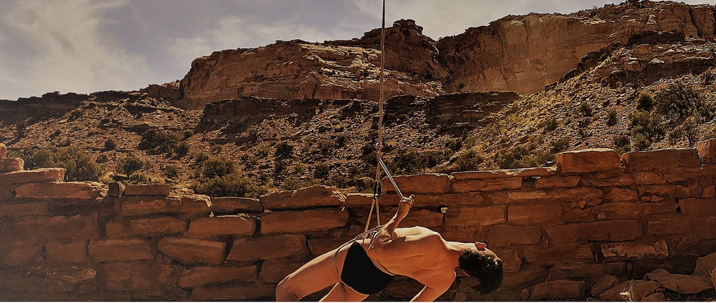 A person in briefs suspended at the waist by a rope, in a rocky desert.