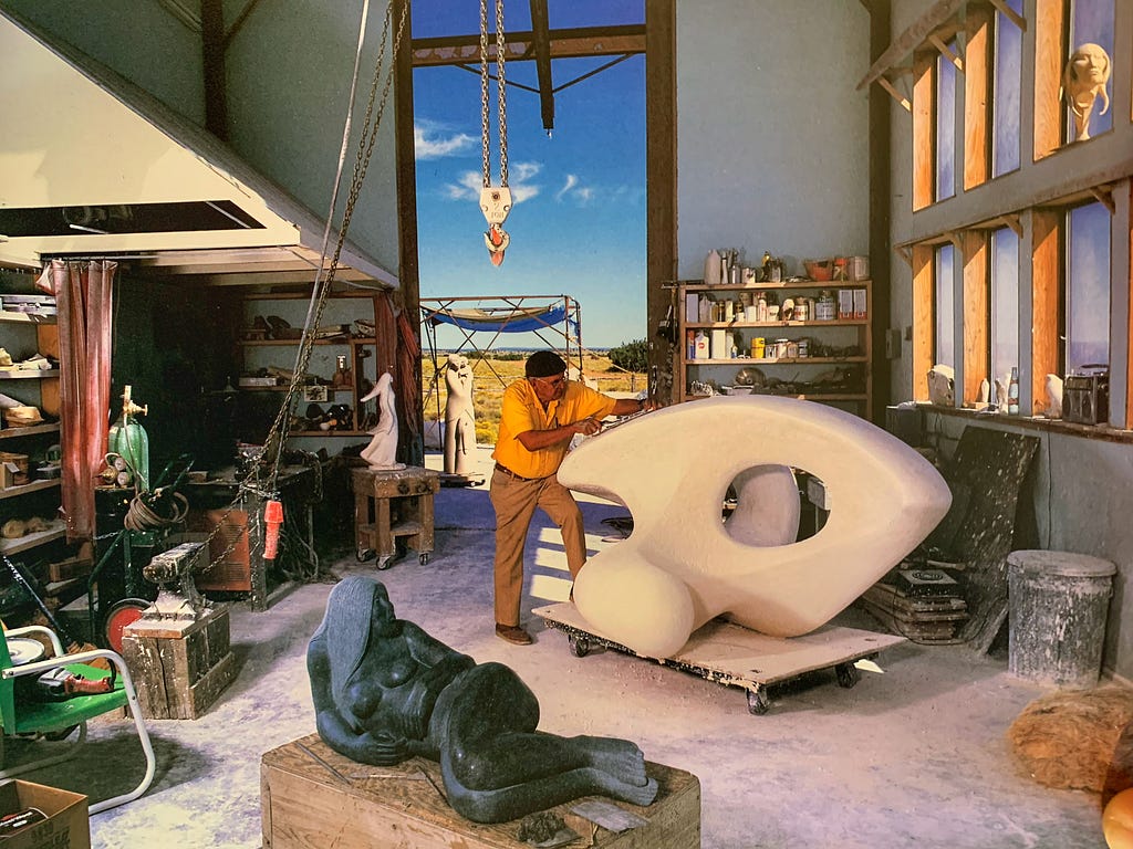 Surrounded by sculpture in various materials, including figures in various poses, a sculptor shapes a large abstract piece. Behind him, shining through a large open doorway, is the blue sky.