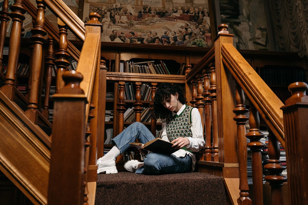 A dark haired person sitting on stairs reading an antique book.