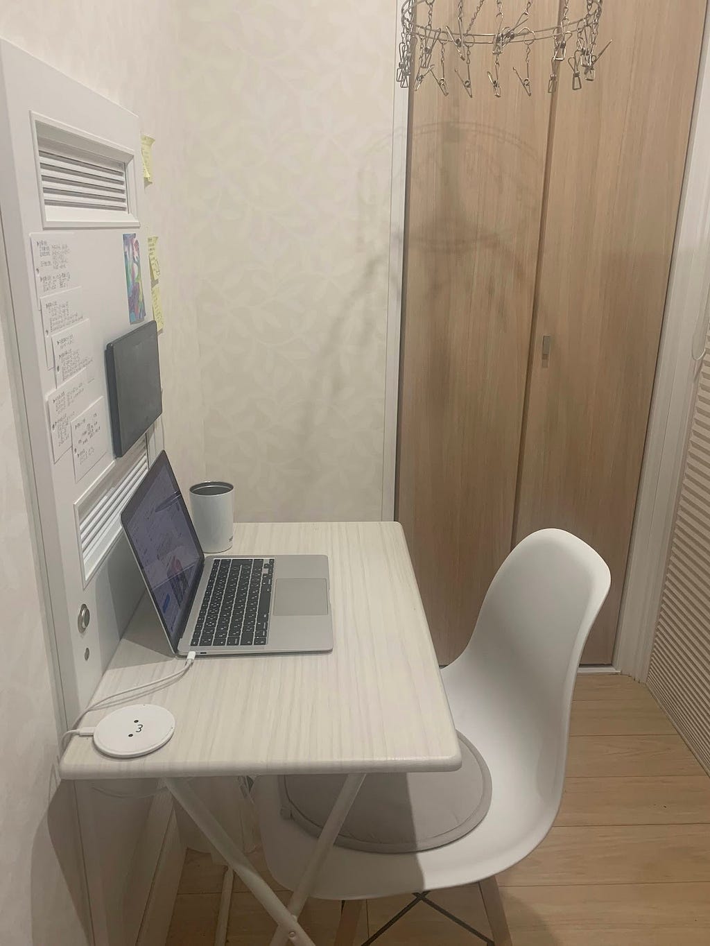 A minimalist writing setup in the corner of a closet, featuring a small white desk with a laptop, a coffee cup, and a white chair. The desk is positioned against a wall with a few notes attached to it and a door of a wooden closet nearby.