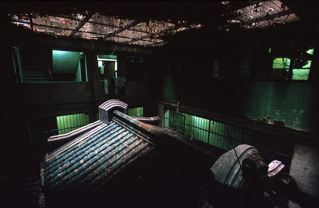 A photograph by Greg Girard showing a temple inside Kowloon Walled City, Hong Kong, viewed from above. The traditional curved ceramic roof tiles of the temple structure are visible in the foreground, now enclosed within and overshadowed by the surrounding concrete building. Eerie green light filters through barred windows and a wire mesh ceiling, illuminating peeling walls, debris, and a staircase in the background. Laundry hangs in a dimly lit upper room.