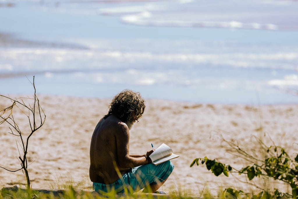 An writer writing on a notebook sitting on a beach alone.