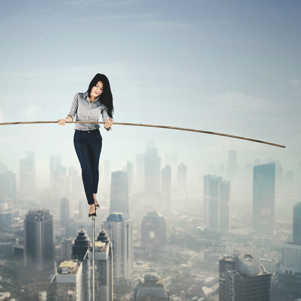 Woman with dark hair in a buttondown shirt, black slacks and high heels walking a tightrope above a smoggy city