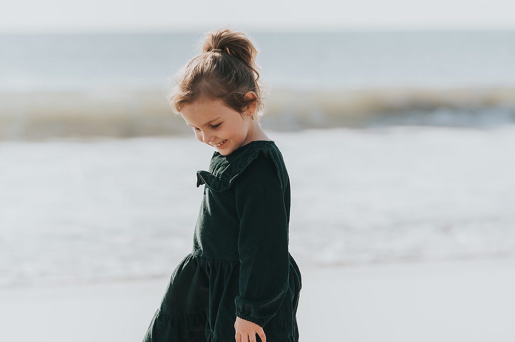 A young child (girl) standing on a beach smiling and looking down at the sand.