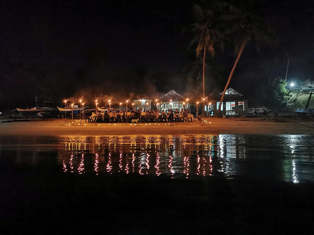 An evening photo of a group of people sitting on a long table on the beachfront eating dinner surrounded by tiki torches.