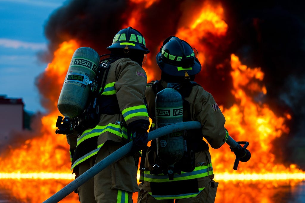 A pair of firefighters facing huge orange flames with a fire hose, photographed from behind.