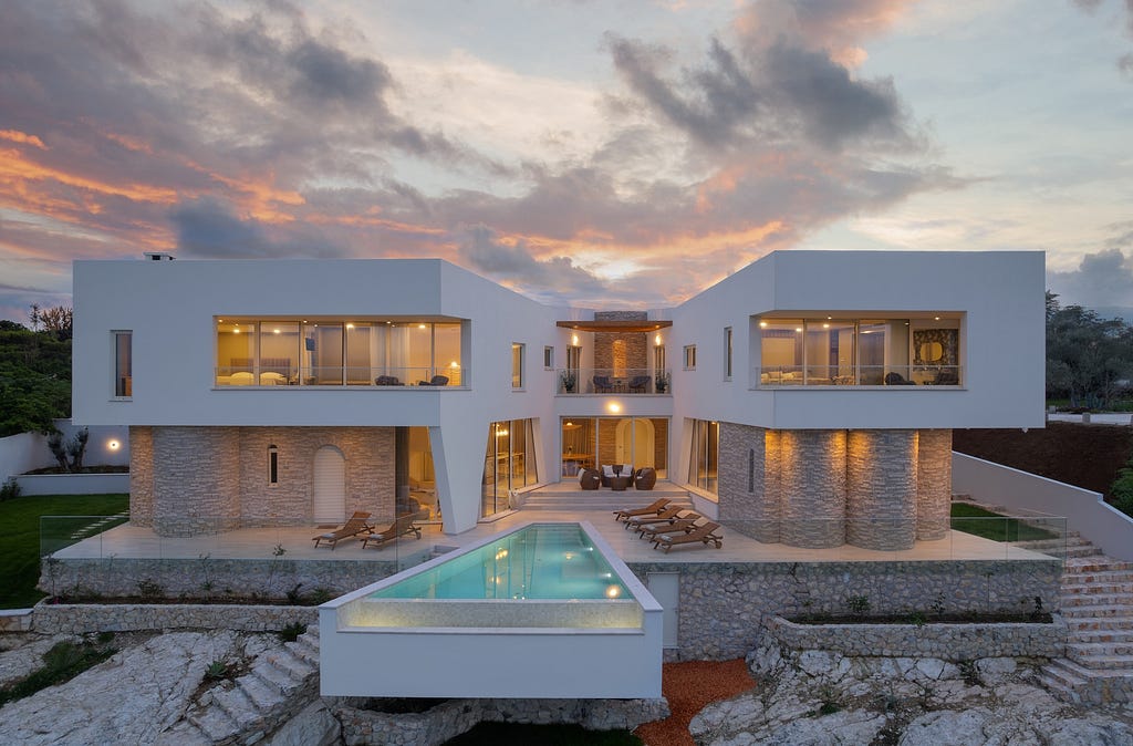 A stunning modern villa at dusk, featuring a striking architectural design with clean lines and large glass windows. The villa is composed of two main wings connected by an outdoor terrace and a central courtyard. The exterior showcases a blend of white walls and textured stone accents. An infinity pool extends outward from the central terrace, surrounded by lounge chairs. Warm indoor lighting creates a welcoming ambiance, contrasting with the cool tones of the evening sky.