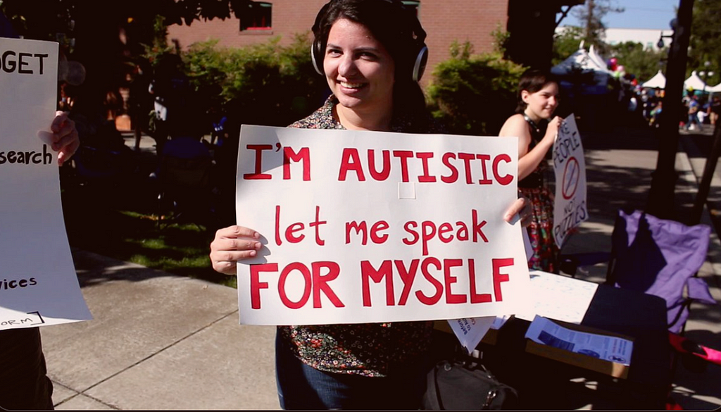 Young white person with noise-cancelling headphones in the street holding a banner that says “I’M AUTISTIC. Let me speak FOR MYSELF.”
