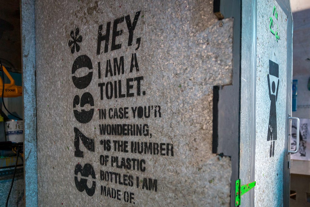 Outside of a portable toilet, made of recyclable materials. Color is silver and black with a graphic of a woman (indicating female toilet) and a bright green recycling symbol on front.