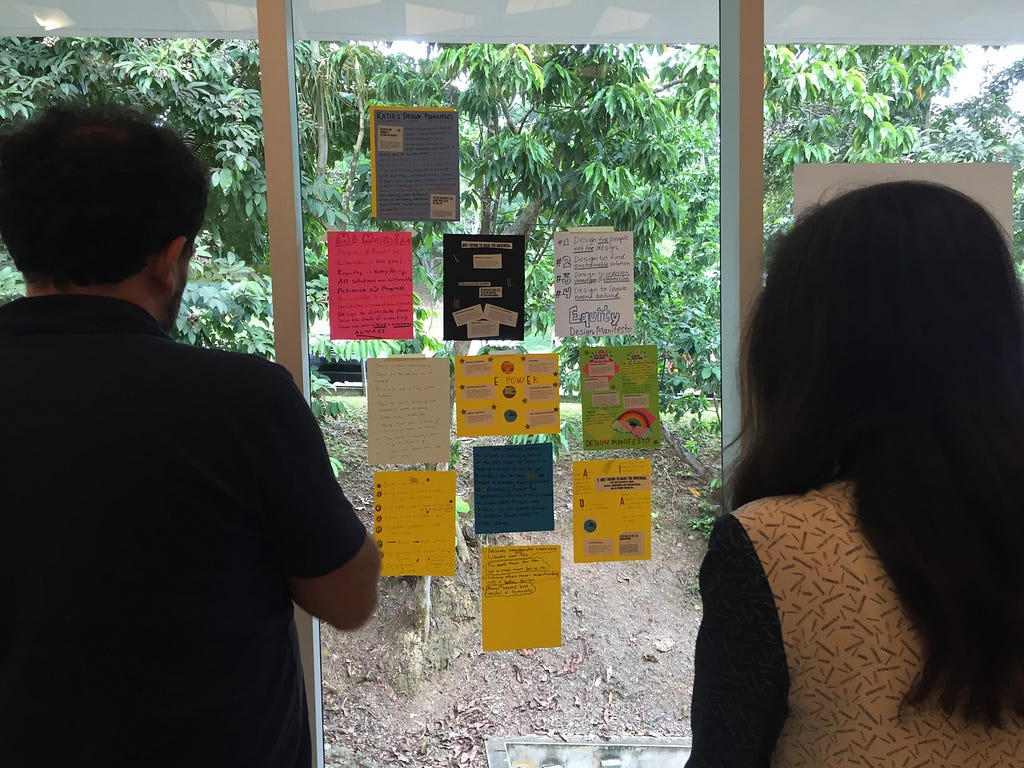 A man and a woman facing a glass wall with posters, taking a closer look at “Design Manifestos” that each participant made.