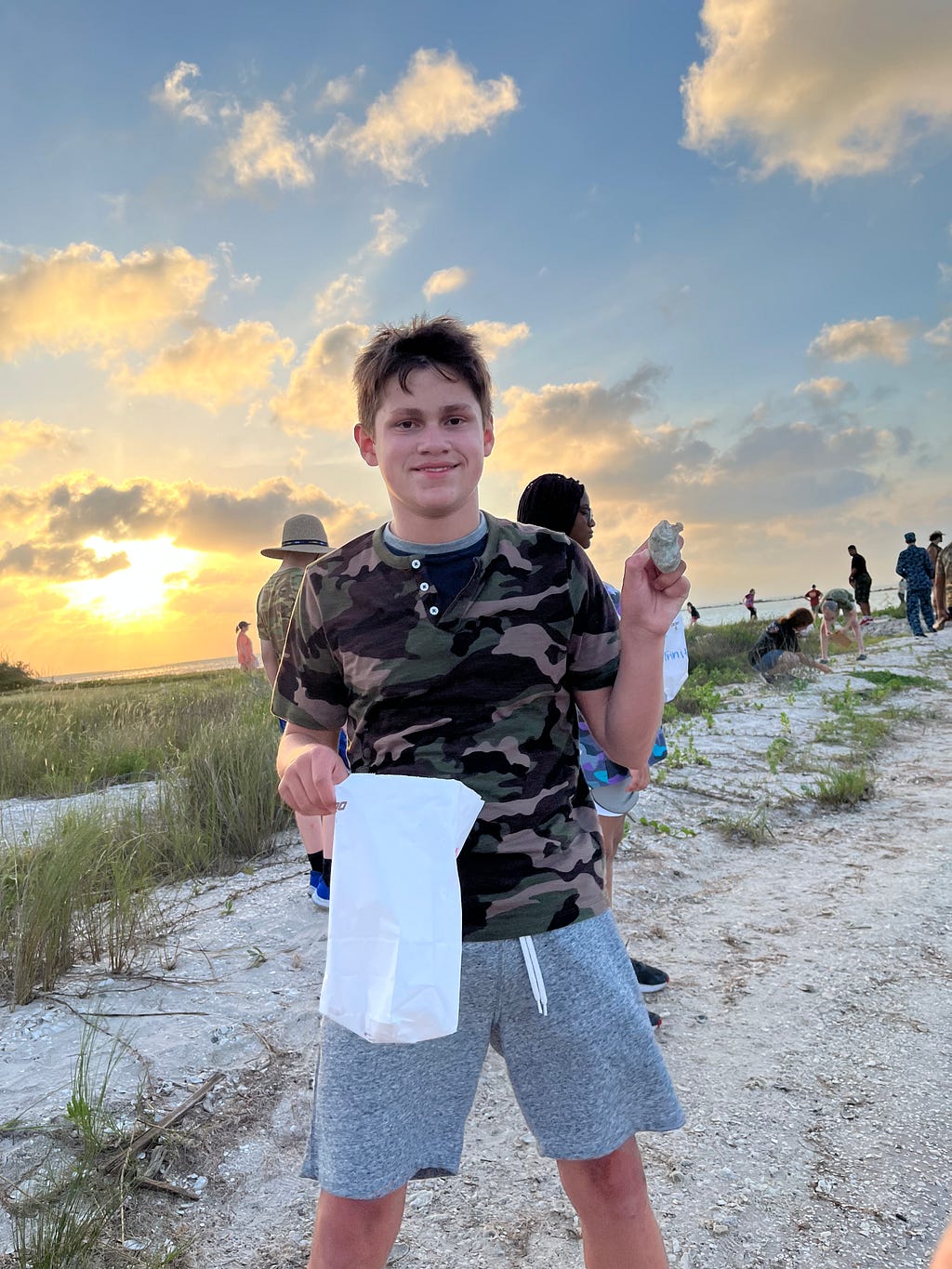 camper holds up shell he found beachcombing on the bay. the rest of the campers are in the background beachcombing. THe sun is setting in the background