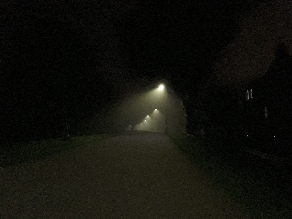 a nightly dark and foggy path in a park, merely suggested by the few lights from the streetlamps guiding through the darkness