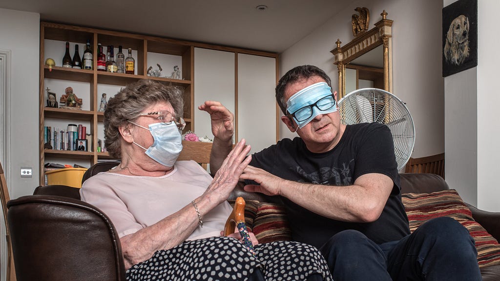 Photograph of a man and his mother sat side by side on a sofa in a living room. The man seated on the right has a medial face mask covering his eyes and is wearing his black rimmed glasses over the top of the mask. He is raising his hands towards his mother as if to try to touch and feel her face. His mother is also wearing a medical face mask but over her mouth and nose and is raising her hand in defence. Rested between her legs is the handle and top half of her walking stick. In the background