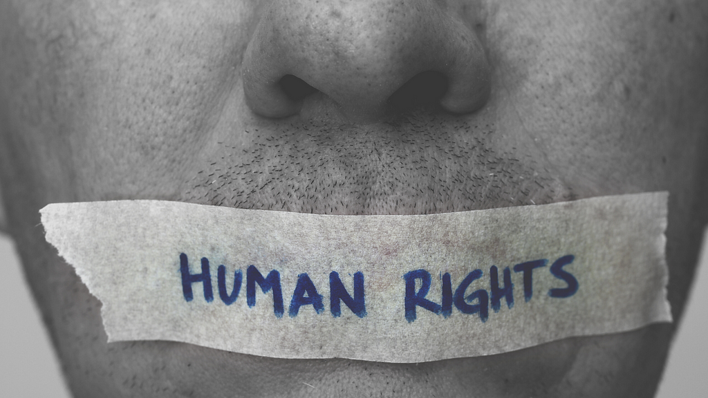 A human face showing the mouth having a piece of paper stuck on it which reads “human rights”
