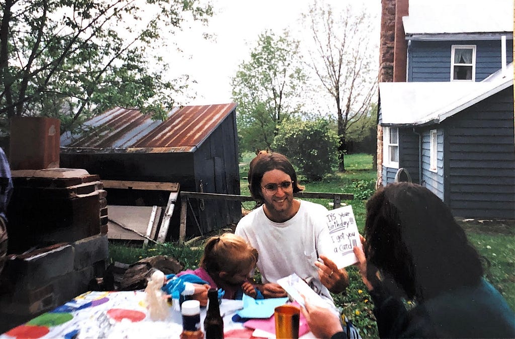 A small child sits outside at a table with her parents. She is leaning against her father, who is smiling. Her mother is in the foreground, facing away from the camera. She is holding a card which reads “It’s your birthday! I got you a card.” In the background there is a shed with a rusted metal roof and a blue house with a chimney.