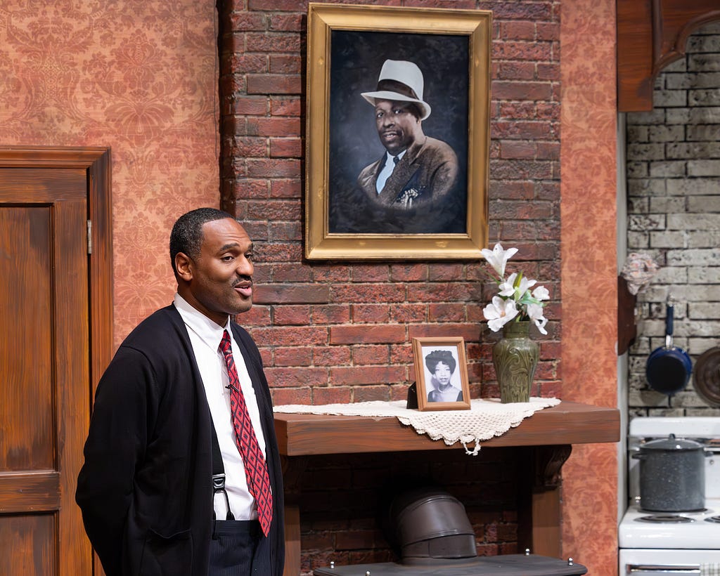 A man in a suit and tie stands in front of a portrait of Father Divine.