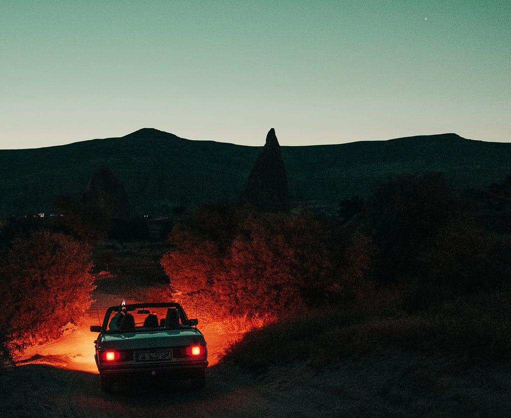Car sitting in a darkened setting, with shrubbery showing to either rear sides, by the glow of the car’s rear brake lights, with only the front parking lights shown. Daylight going off in the upper half of the landscape where the car is located.