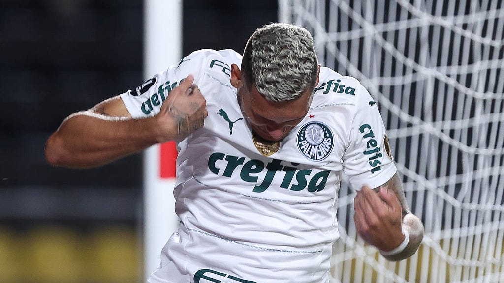Rafael Navarro, atacante do Palmeiras, comemora gol em jogo pela Libertadores. Ele veste camisa branca, com detalhes em verde na horizontal, bem finos, e tem cabelo platinado. Ele faz movimento dos braços de cima para baixo, como quem “espanta a zica”