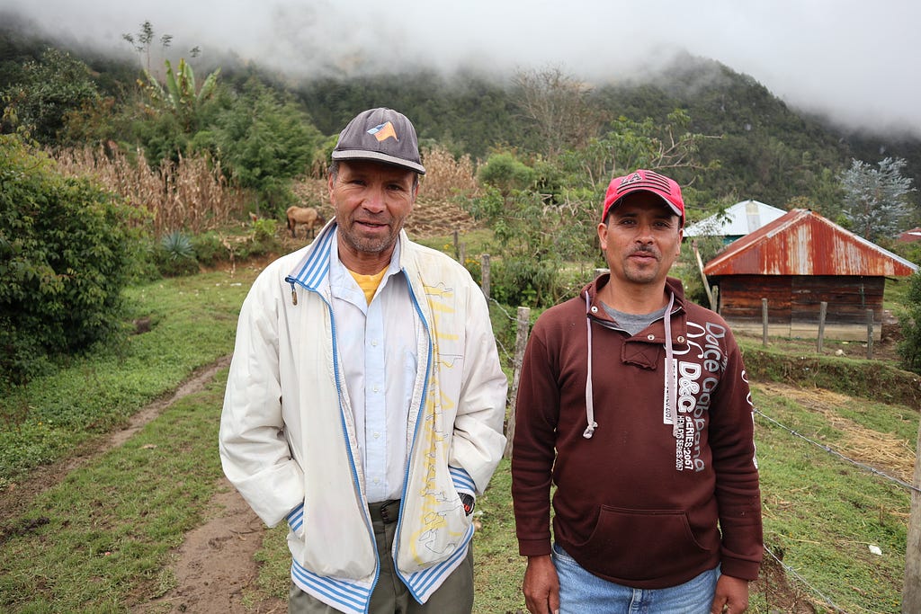 An image of two men in a rural location in Guatemala. They are standing in the foreground. In the background are some buildings in cleared areas among the vegetation. In the far background are hills partially shrouded in mist.