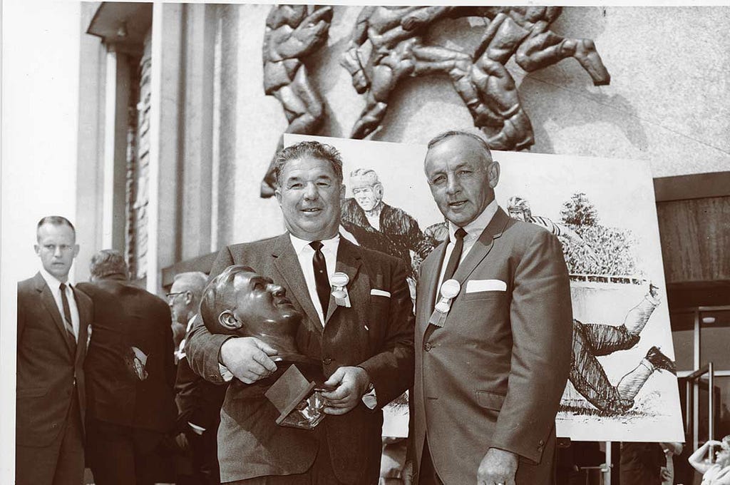 black and white photo of Arnie Huber holding his own bust and posing for a photographi with a man in a suit during Huber’s Hall of Fame induction ceremony.