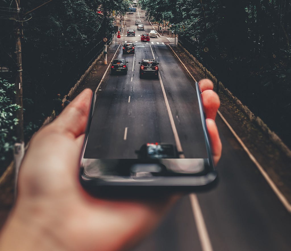 A hand grabs a cellphone from where we see cars leaving to a street, making referrence to web Augmented Reality, or web AR | Adapted photo of Matheus Bertelli, available in Pexels.