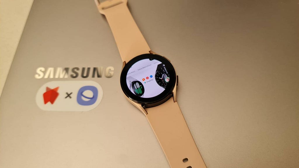 A Samsung Galaxy Watch4 placed on top of a Samsung Laptop. The watch shows the Samsung homepage on it’s face.