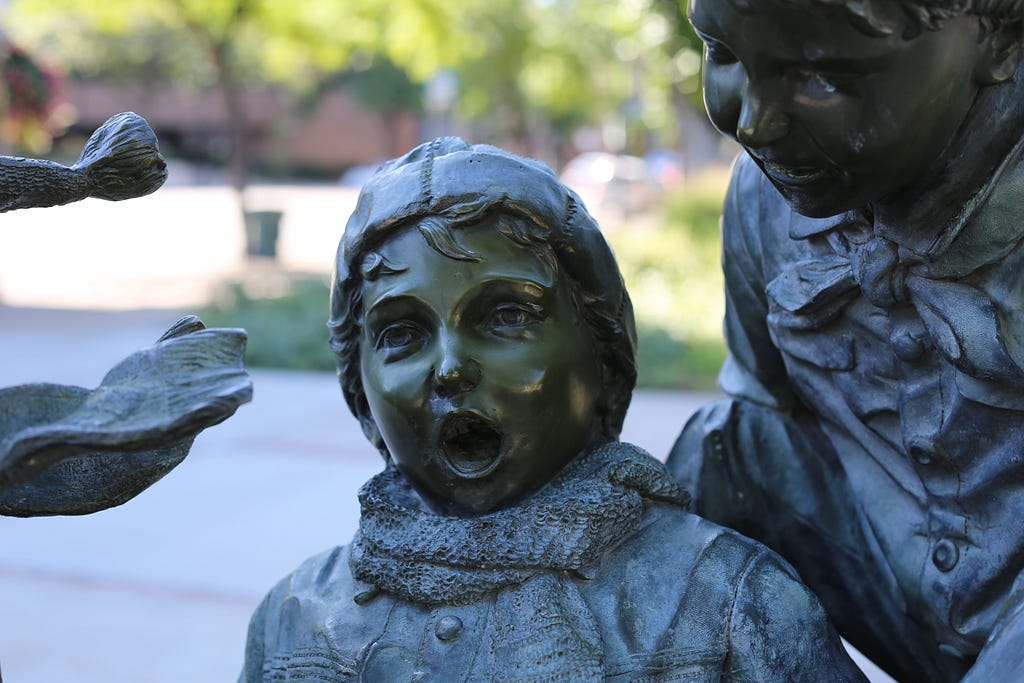 Outdoor bronze statue with a child feigning surprise and another child trying to tell them something.